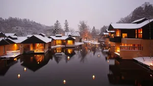 Snow-covered waterfront houses with warm interior lights reflected in calm water.