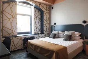 A modern, minimalist bedroom with geometric patterns on the wall and a bed with white and brown linens, under a large window.