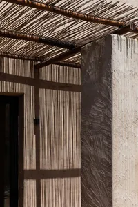 Concrete columns casting shadows on a wall, with a grass and wood roof structure.