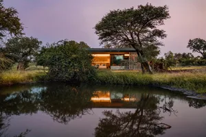 A modern house with a glass and metal exterior sits by a serene water body, surrounded by lush greenery.