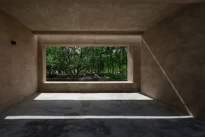 Modern interior space with concrete walls and floor, framed by a large window showcasing a green outdoor area with trees.