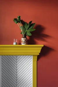 A modern mantle with a green plant and a colorful vase on top, against a red wall.