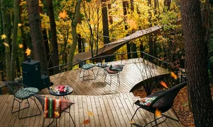 A deck surrounded by autumn foliage with wooden furniture and a grill.