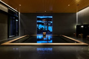 Indoor spa with a rectangular hot tub, made of wood and concrete, surrounded by dark stone walls and floor, illuminated by so