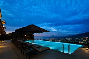 A modern poolside area with concrete walkways and illuminated pool at night, overlooking a cityscape under a dark, cloudy sky