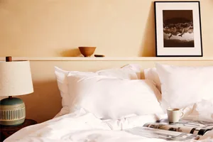 A modern bedroom featuring a neatly made white bed with various pillows, a nightstand with a lamp, and a coffee mug on a maga