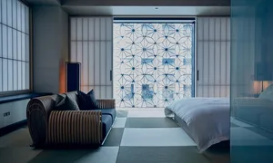 A minimalist hotel bedroom with a wooden sofa, a bed with white linens, and a sliding door with a geometric pattern.