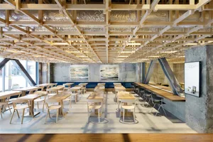 A modern cafe interior featuring wooden tables and chairs, exposed wooden beams, and concrete walls and flooring.