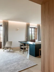 Modern and minimalistic office space featuring a blue desk, wooden chair, and fabric curtains.