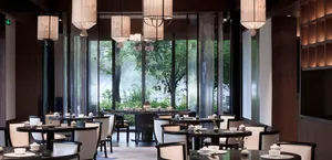 A modern restaurant interior with elegant lighting fixtures and large windows showcasing greenery.