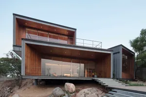 A modern, two-story house with industrial design features, including wood siding, large glass windows, and concrete steps.