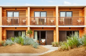 A two-story vacation home with a wooden exterior, outdoor seating areas, and sandy surroundings.