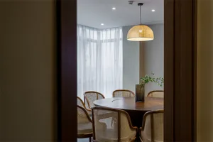 A modern dining room featuring wood furniture, large windows with sheer curtains, and a woven pendant light hanging above the