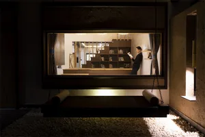 A man reads a book on a floating platform in an industrial-style living space with a bookshelf and wooden elements.