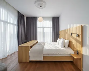 Modern minimalist bedroom with a wooden floor, bed, and chairs, featuring a light pendant chandelier and large windows with s