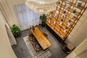 A minimalist living room with a wooden table, chairs, and bookshelf, featuring light-colored textiles and plants.