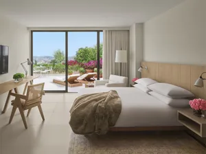 Modern bedroom with white walls, glass door to balcony, wooden bed frame, white bedding, and beige blanket.