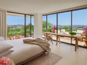 Modern bedroom with balcony overlooking cityscape, featuring a wooden bed frame, glass window, and fabric textiles.