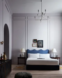 A modern minimalist bedroom with a blue upholstered headboard, wooden flooring, and framed art on the wall.
