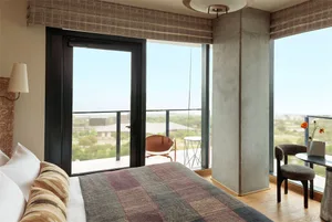 A modern bedroom featuring concrete flooring, a wooden bed frame, and floor-to-ceiling windows overlooking a cityscape.