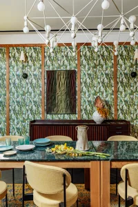Modern dining room with wooden and marble furniture, featuring a white geometric chandelier and green and brown botanical wal