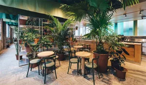 Interior view of a cafe with round wooden tables, black chairs, and various potted plants.