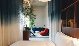 A modern bedroom featuring a bed with white sheets and pillows, a blue and red armchair, a green plant in a brown planter, an