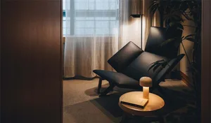 A modern living room with a black leather lounge chair, fabric cushions, and a wooden side table with a book and lamp.