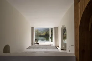 Minimalist bedroom with concrete walls and floor, framed by large windows overlooking a scenic lake view.