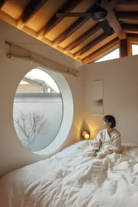 A person wearing a white robe sitting on a bed with white linens, looking out of a circular window