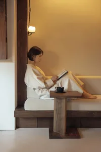 A minimalist living room features a wood sofa with a small table and a woman reading a book and drinking coffee.