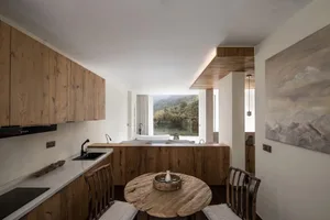 Modern minimalist kitchen with wooden cabinets and table, featuring a view of a serene landscape through a large window.