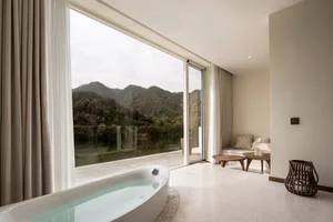 A modern bathroom featuring white marble flooring, glass walls with a view of the landscape, and a concrete sofa in the adjac