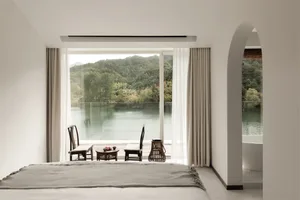 A modern, minimalist bedroom featuring white walls, wood furniture, and a glass window overlooking a scenic lake and mountain
