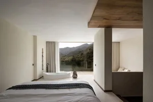 A modern bedroom with a concrete floor, a white bed with a black blanket, and a freestanding bathtub overlooking a scenic mou