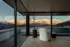 A minimalist balcony with wooden flooring, featuring a white chair and a tree pot, overlooking picturesque snow-capped mounta