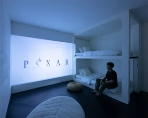 A minimalist living room featuring white walls, floor, and furniture, with a person sitting on a built-in bed under a Pixar l