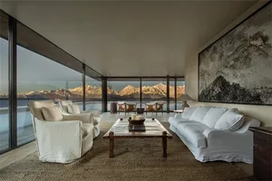 A modern, minimalist living room with a white sofa, armchairs, and a wooden coffee table featuring candles and a bowl. Large 