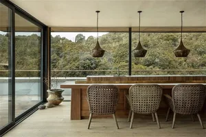 A modern kitchen with a wooden counter, wicker chairs, and large windows overlooking a forested area.