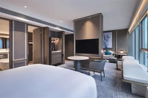 A modern bedroom with a neutral color palette featuring a large bed, a seating area, and a large window.
