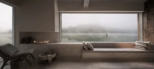 A modern living room with a concrete fireplace, large window overlooking a serene lake, and a person paddleboarding on the fo