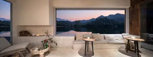 Modern, minimalist living room with concrete walls, large windows overlooking a serene lake and mountains at dusk.