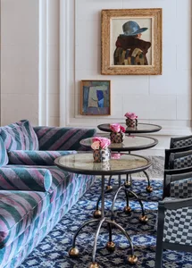 A modern lounge featuring a colorful velvet sofa, glass side tables with flowers, and a patterned rug.