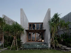 A modern vacation home with a concrete exterior, surrounded by lush greenery and palm trees, overlooking the ocean at sunset.