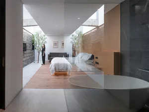 A minimalist bedroom featuring a white bed and wooden flooring, with a reflection in a glass wall.