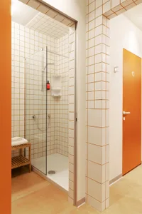 A minimalist bathroom with white tiles, glass shower enclosure, and orange accents.