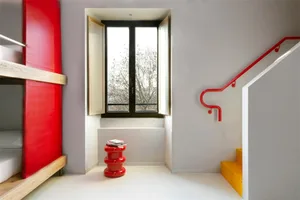 A minimalist bedroom with red, white, and yellow accents, featuring open windows and a red stool.