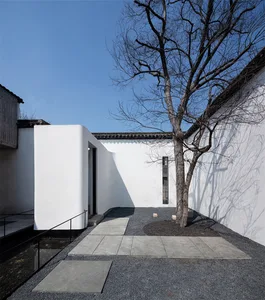 A minimalist white-walled courtyard with concrete pathways and gravel, featuring a leafless tree and traditional architectura