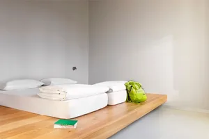 A modern minimalist bedroom with white bedding, a wooden floor, and a green backpack.