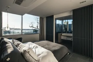 A modern minimalist bedroom featuring a bed with white and gray bedding, a large window with views of the outdoors, and a bat